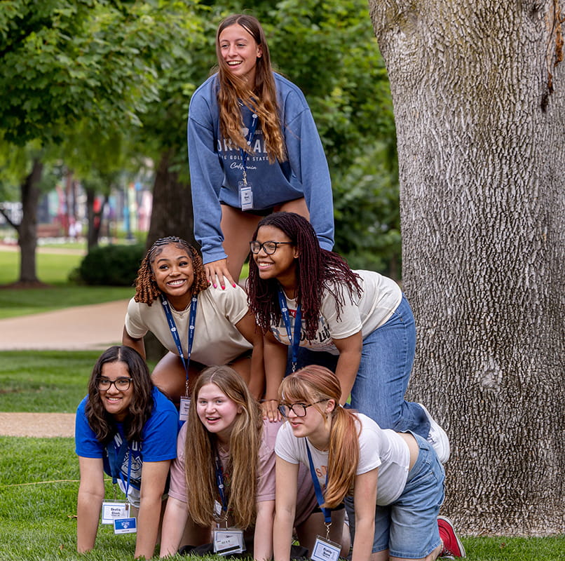 A group of students form a pyramid outside under a tree.