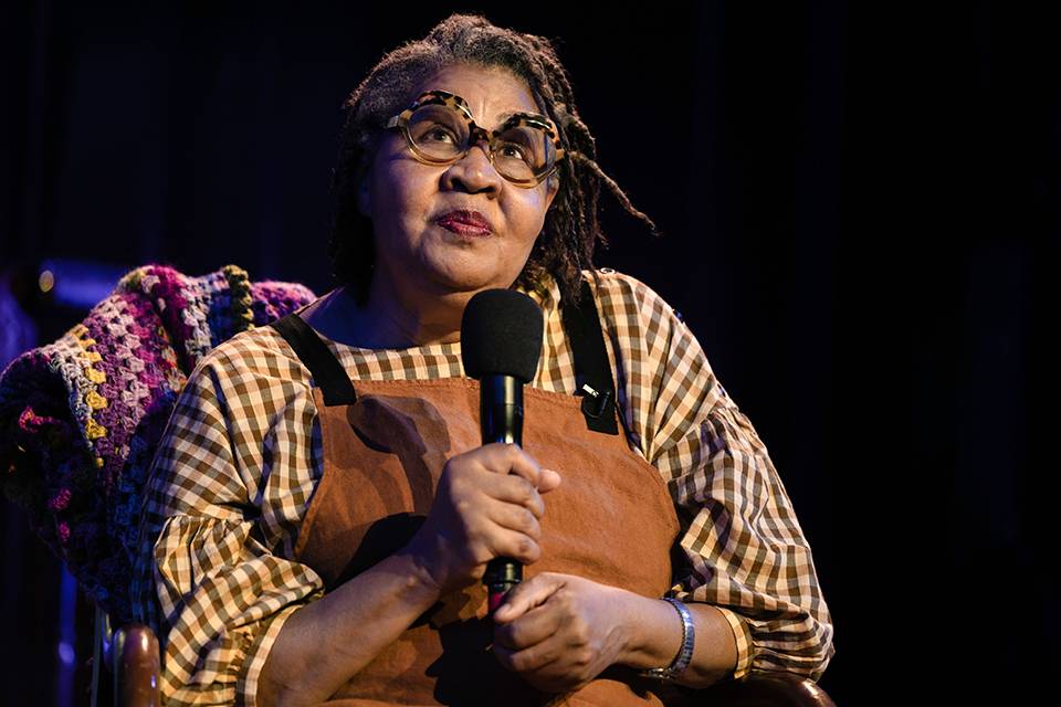 Author Jamaica Kincaid holds a microphone while sitting on a darkened stage.