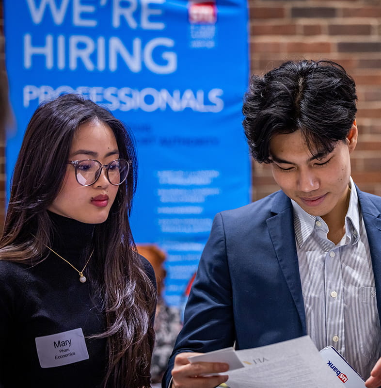 Two students look at a piece of paper while standing in front of a banner that says We're Hiring Professionals.