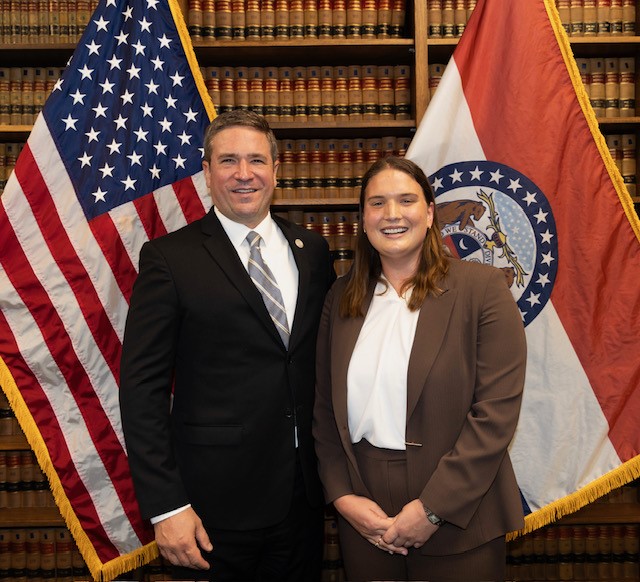 Paige Mitchell stands on the right with Missouri Attorney General Andrew Bailey on the left. They stand in front of a bookcase between the Missouri State flag and the United States flag Paige Mitchell stands next to Missouri Attorney General Andrew Bailey between the Missouri State flag and the United States flag
