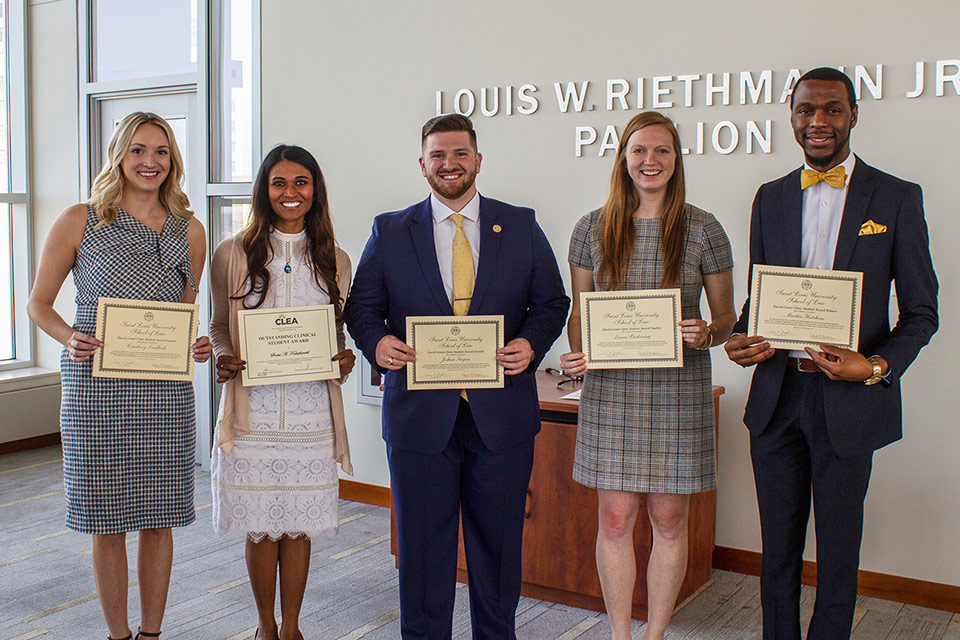 Five students wearing formal attire stand in a line. They are holding yellow certifcates for the 2019 CLEA and David Grant finalists awards. They are standing in front of a wall that have silver words spelling out 'Louis W. Riethman Jr'. The students' head are in front of some of the letters.