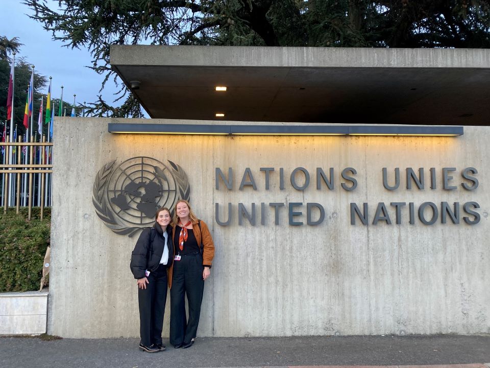 Two students stand outside a building with the words Nations Unies, United Nations. There are international flags and trees in the background.