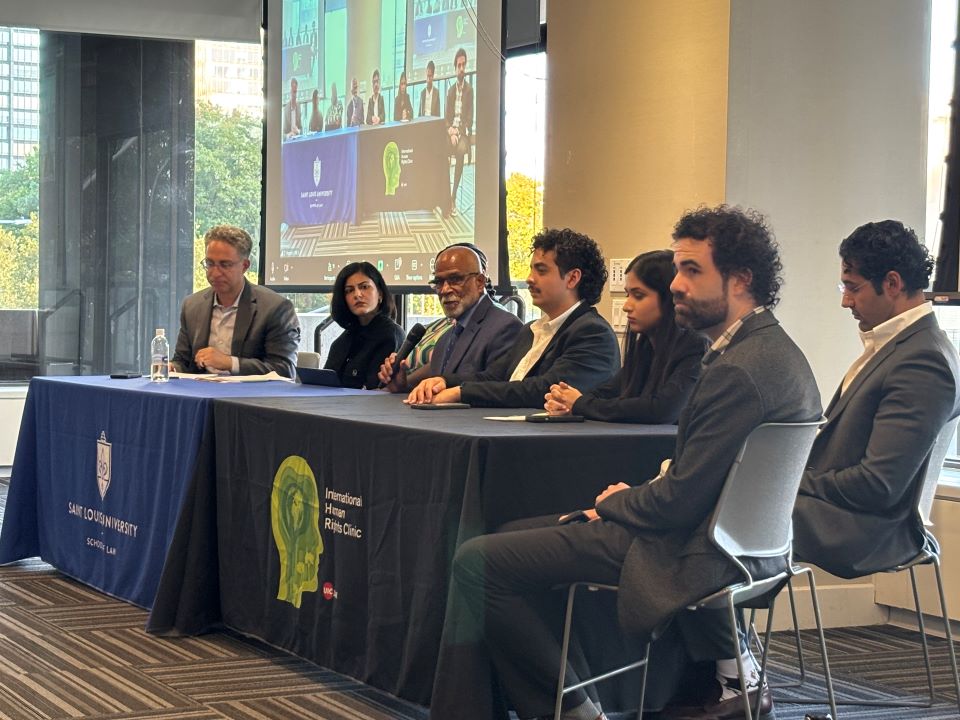 representatives for human rights organizations sit at a table during a press conference at the united nations in new york city for the people's upr Panelists sit at two long tables; one draped with a SLU Law table cloth, the other with a drape from the International Human Rights Clinic.