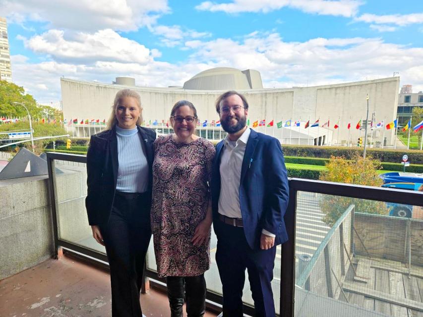 Professor Bartlett, center, poses with students outside the United Nations building with multiple international flags fly in the foreground. Professor Bartlett poses with students outside the United Nations building in New York