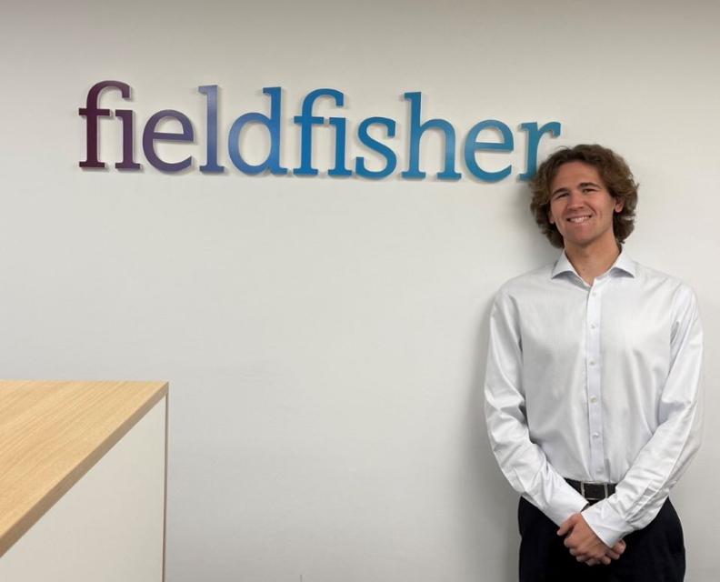 A student posers for a photo next to a wall sign that reads Field Fisher.