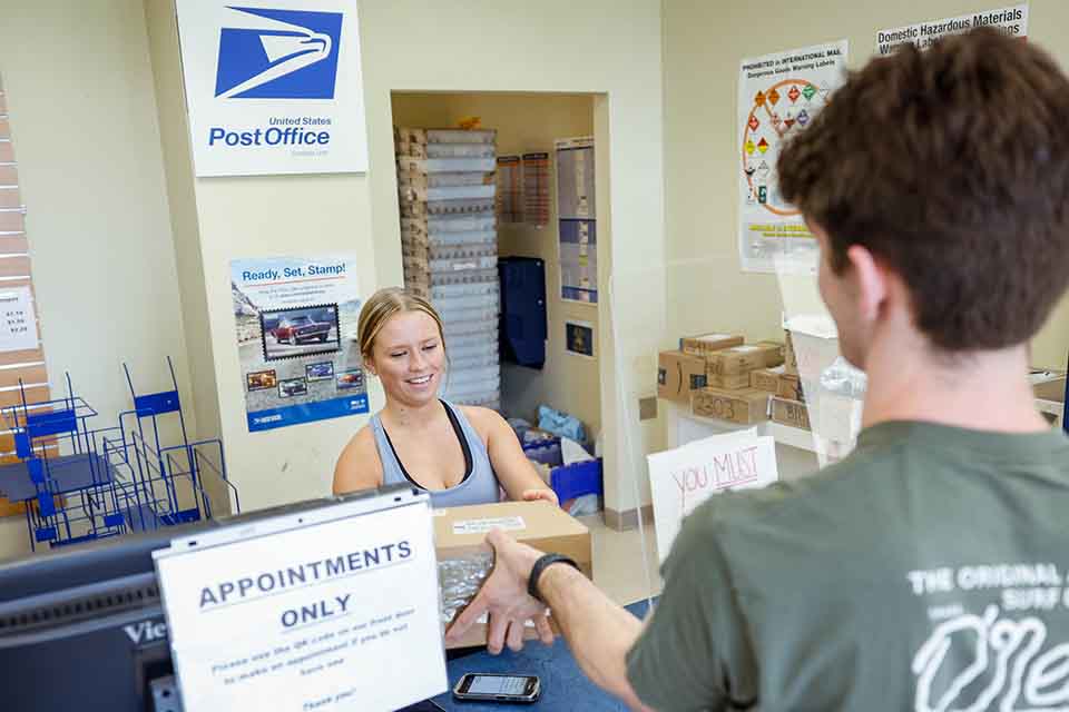 A mailroom worker surrounded by packages hands a student (seen from the back) his mail