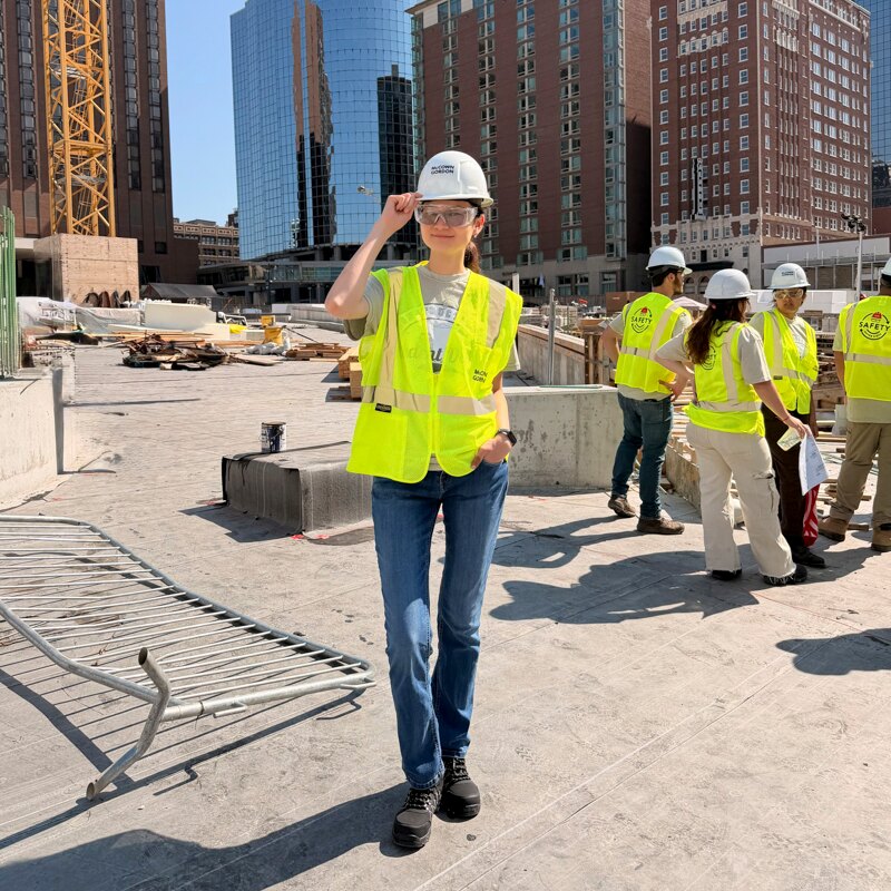 Ayana standing at a construction site wearing a hard hat and a reflective safety vest. 