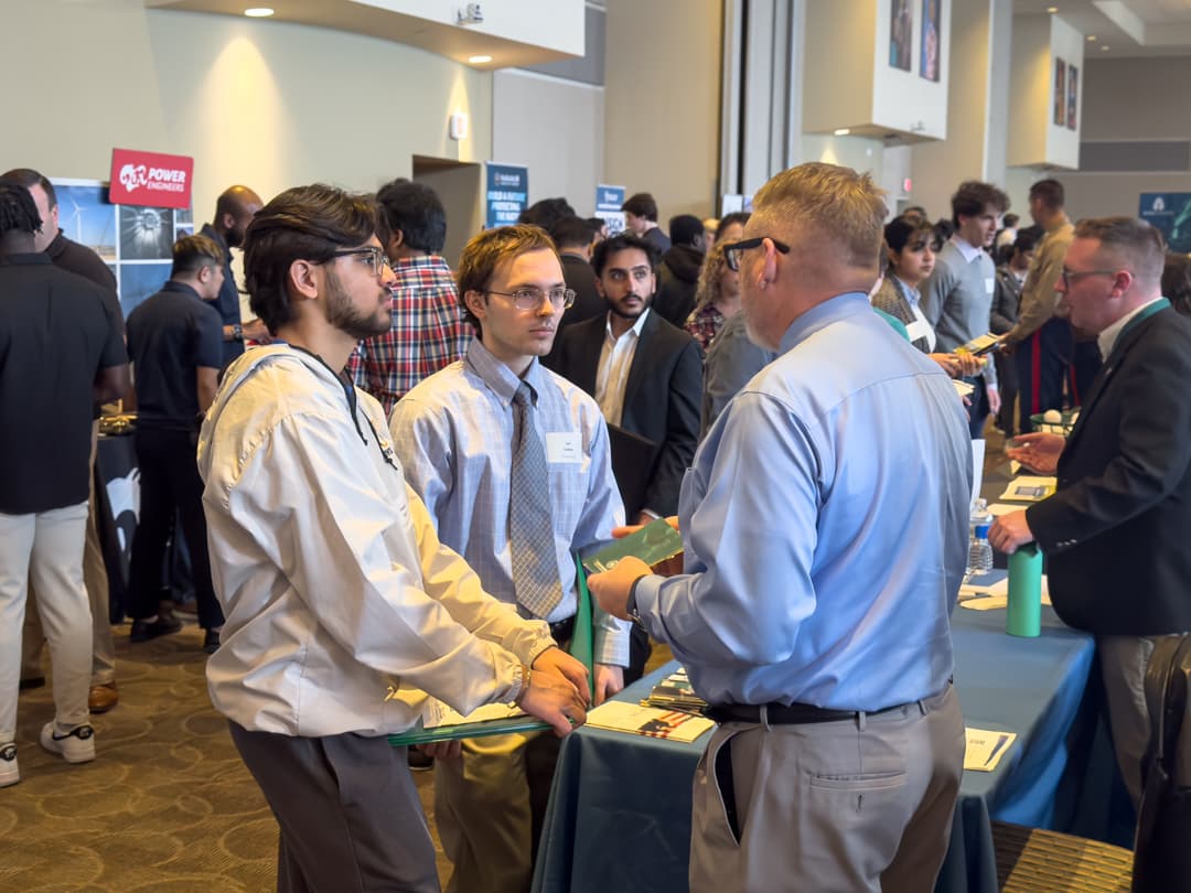 Tw students talk to a prospective employer in a room filled with booths and people.
