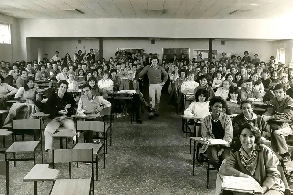 A black and white photo features students sitting at classroom desks while a professor stands in the center of the room.