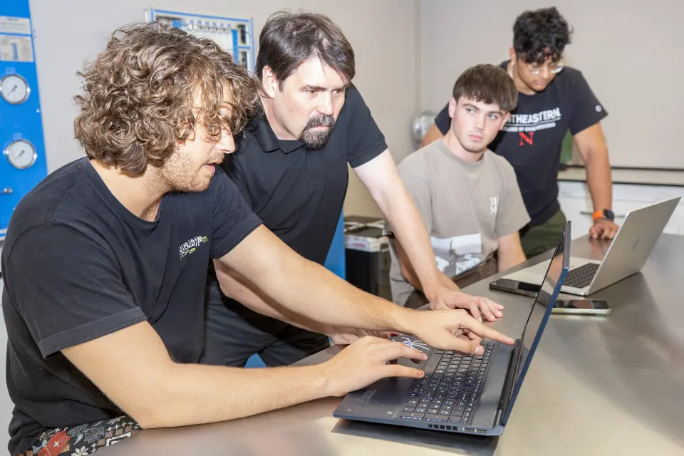 Two students and an instructor examine a computer screen while having a discussion.