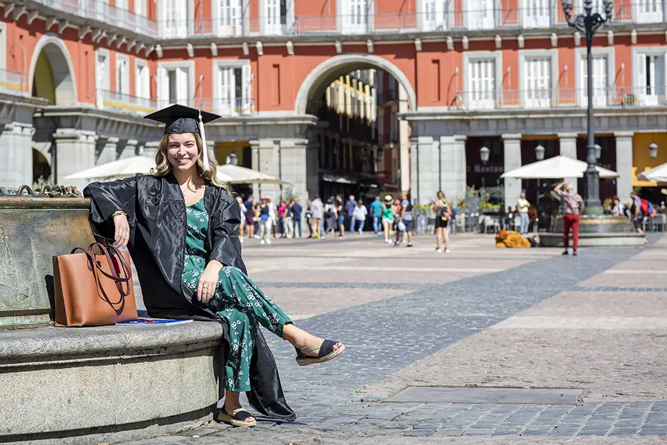 Honors Program student in graduation cap and gown with honors cords sits on a bench in surrounded by brick buildings in Madrid's Plaza Mayor.