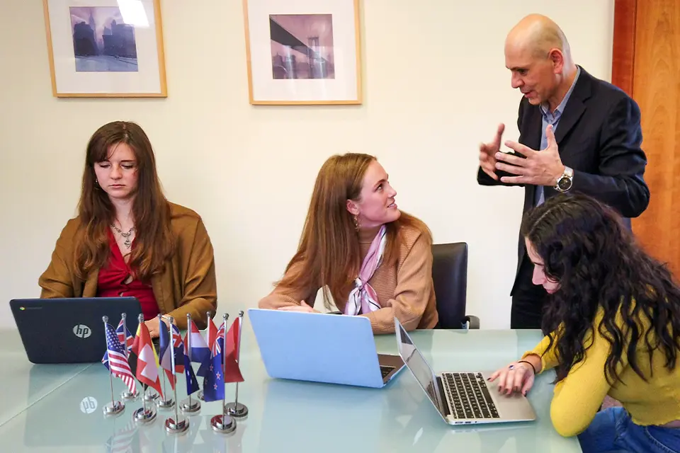 Three students talk with a professor while they sit at a table with laptops and miniature international flags.