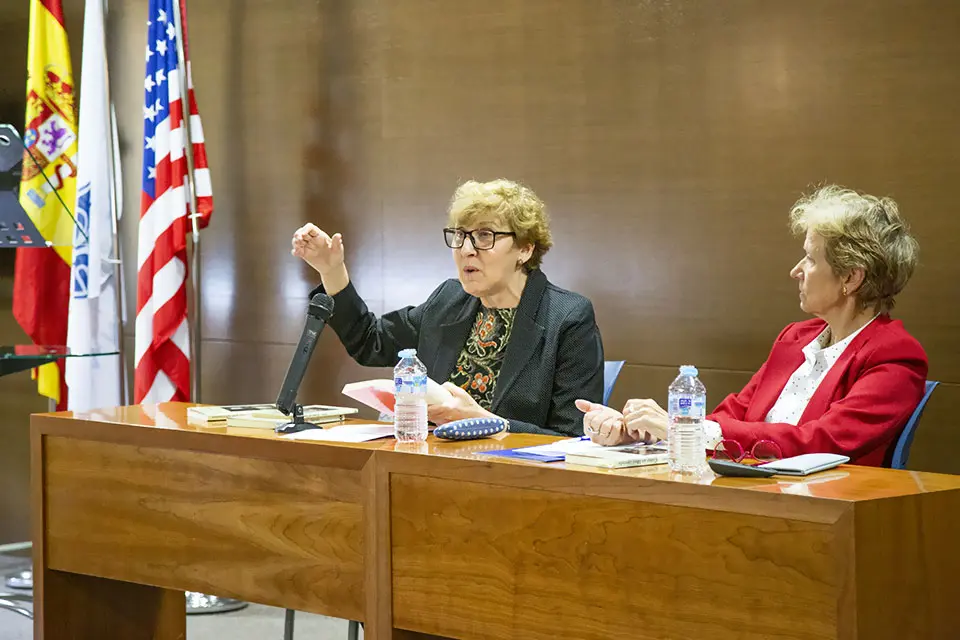 Two women sitting at a table and debating with 3 flags behind them