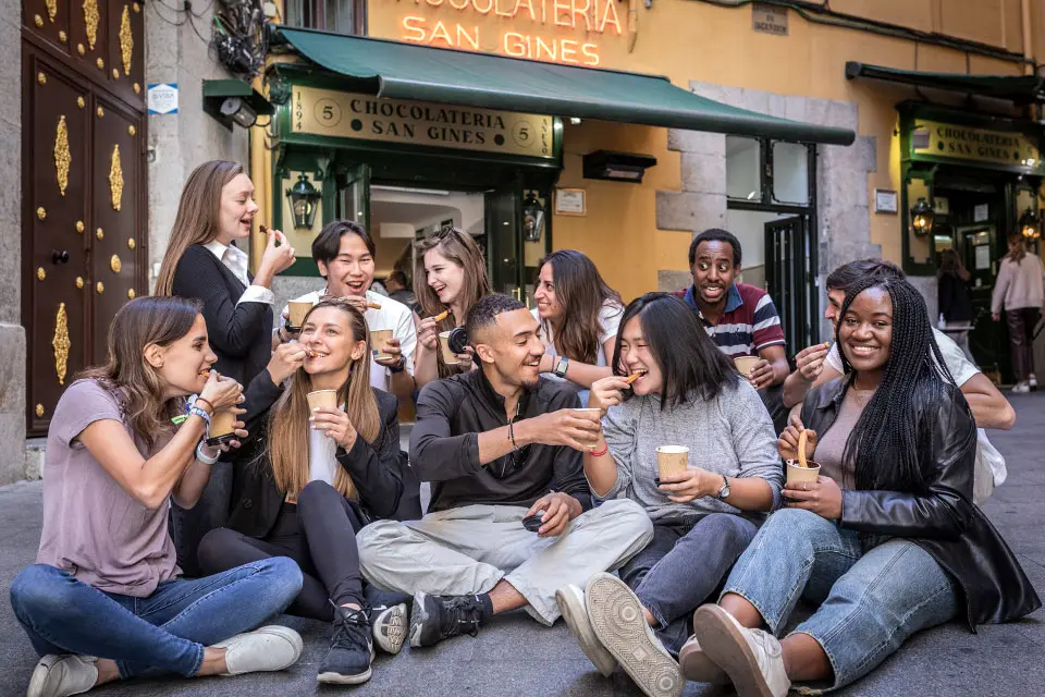 Students sit in front of a store called Chocolateria San Gines dunking pastries in cups of hot chocolate and eating them.
