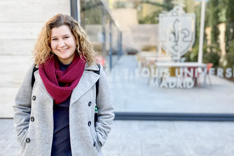 A student stands outside of a SLU Madrid building, wearing a coat, scarf and backpack.