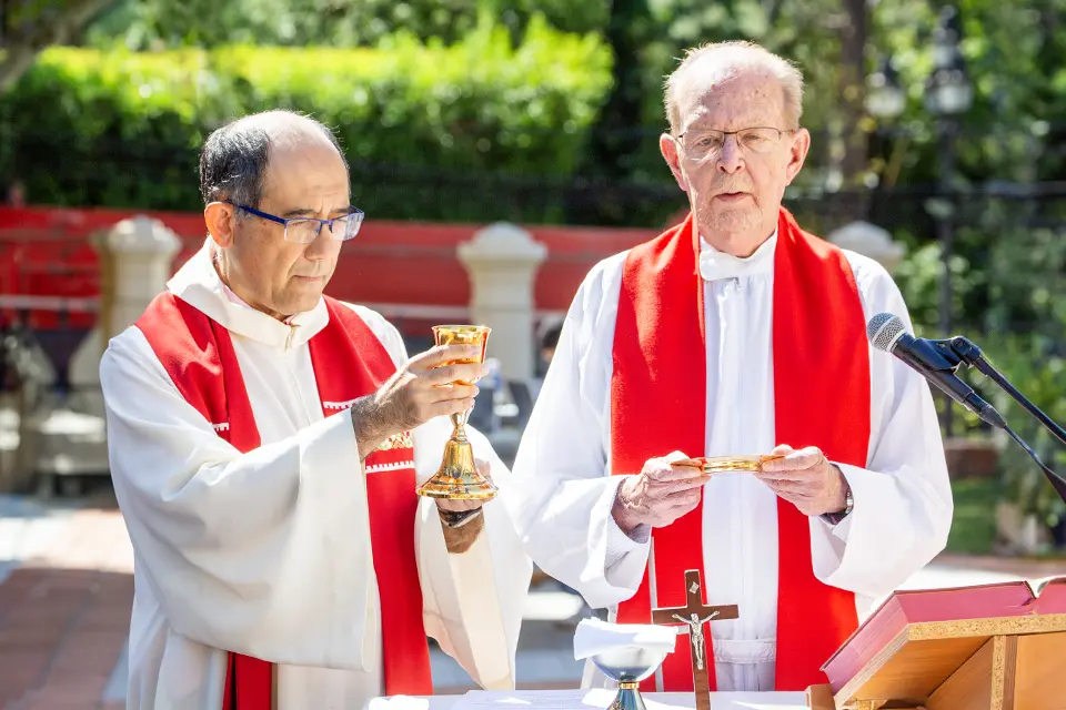 Students line up in an outdoor area while a priest marks a student's forehead. A choir of students stands nearby holding black choir books and singing.