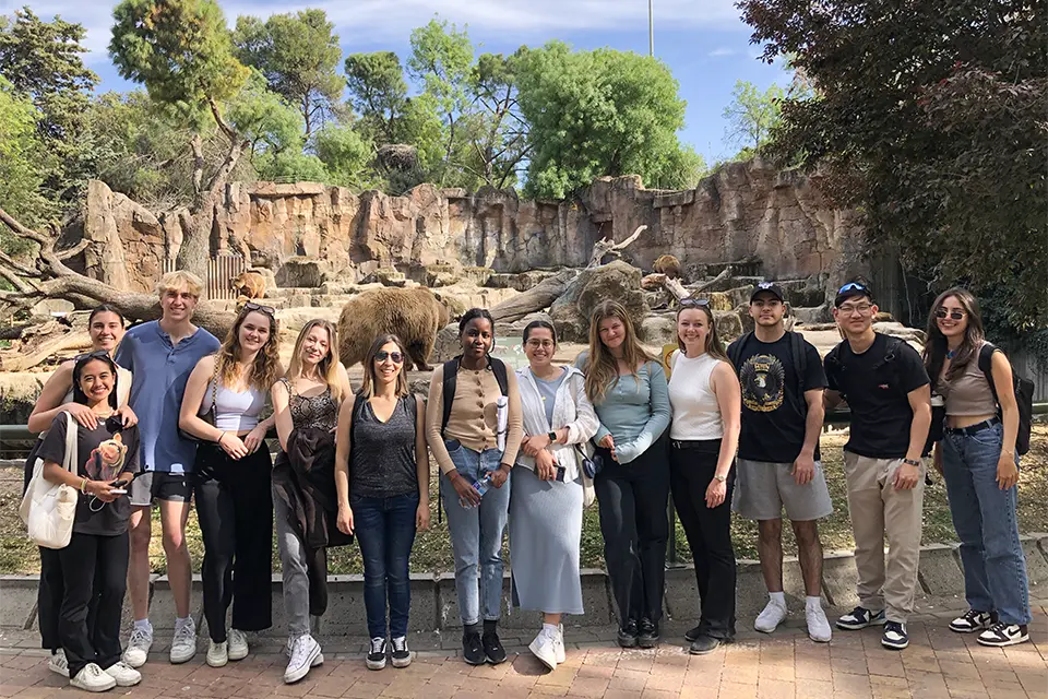 Students and professor stand in front of a rocky animal habitat with bears in the backround.l