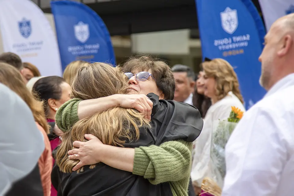 A woman hugs a participant in a graduation ceremony and looks up at the sky, while others mill around the area.