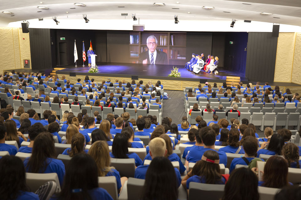 Convocation of the Class of 2026 at Pablo VI Auditorium. Students, most wearing blue T-shirts, face the stage with a video of President Fred Pestello speaking.