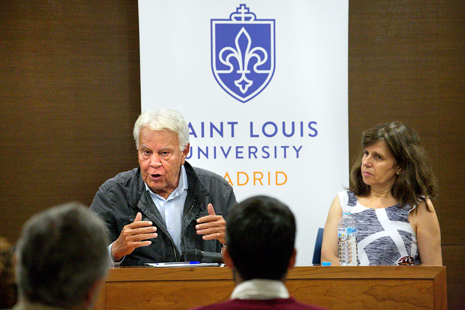 Dr. Laura Tedesco sits next to former Spanish Prime Minister Felipe González during a discussion. They sit at a wooden table in a lecture hall.
