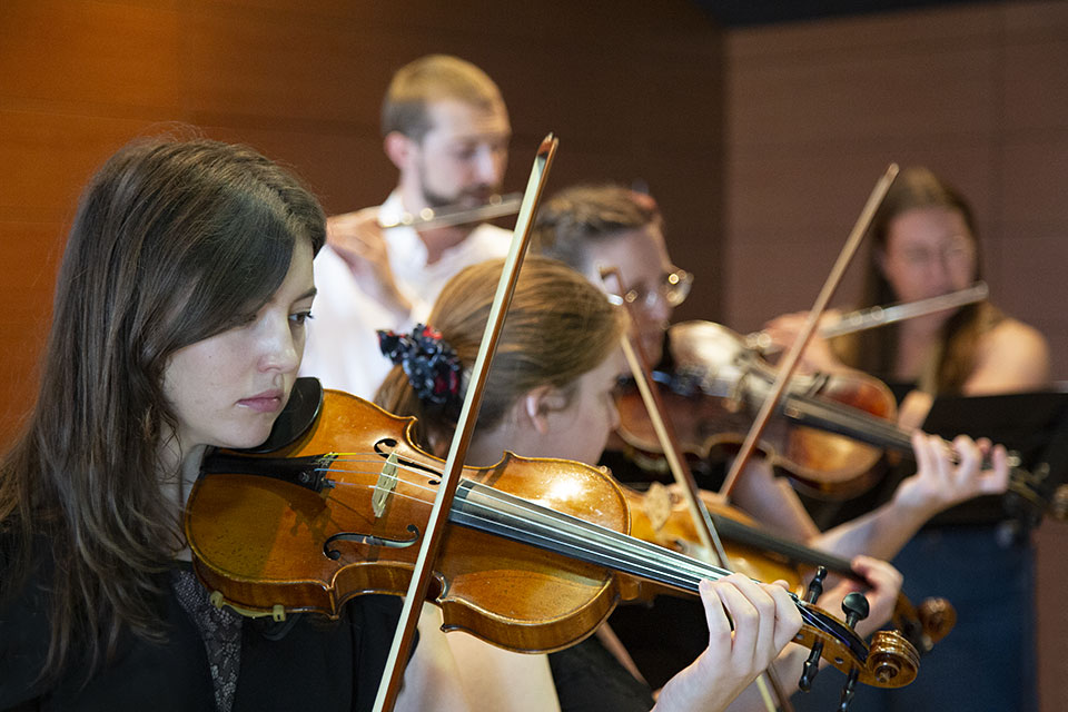Three students play violins while two students play flutes in the background.