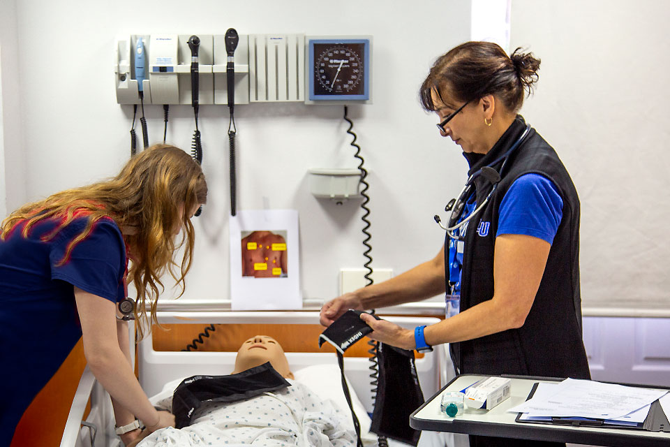 An instructor stands on one side of a hospital bed containing a patient mannequin holding a blood pressure cuff while two students watch from the other side.