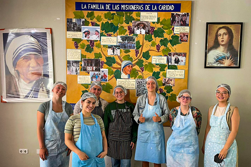 A group of seven students wearing aprons and hairnets pose in front of a large painting that reads La Familia De La Misionera De Caridad.