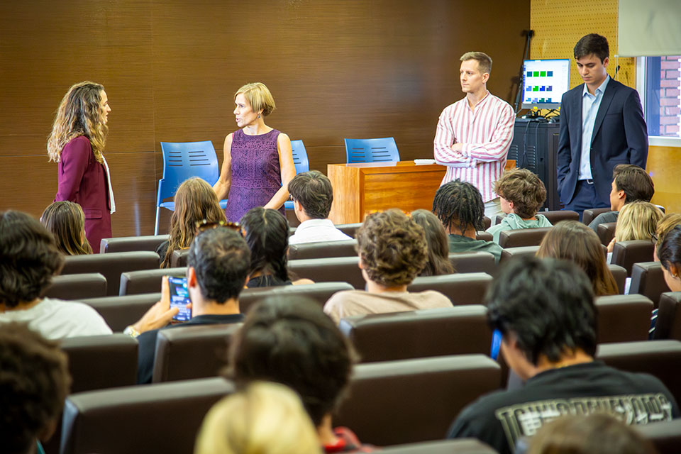 Four presenters share a discussion at the front of an auditorium with students, seen from behind, in the audience.
