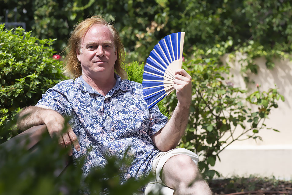 Brian Goss sits in a garden waving a fan.