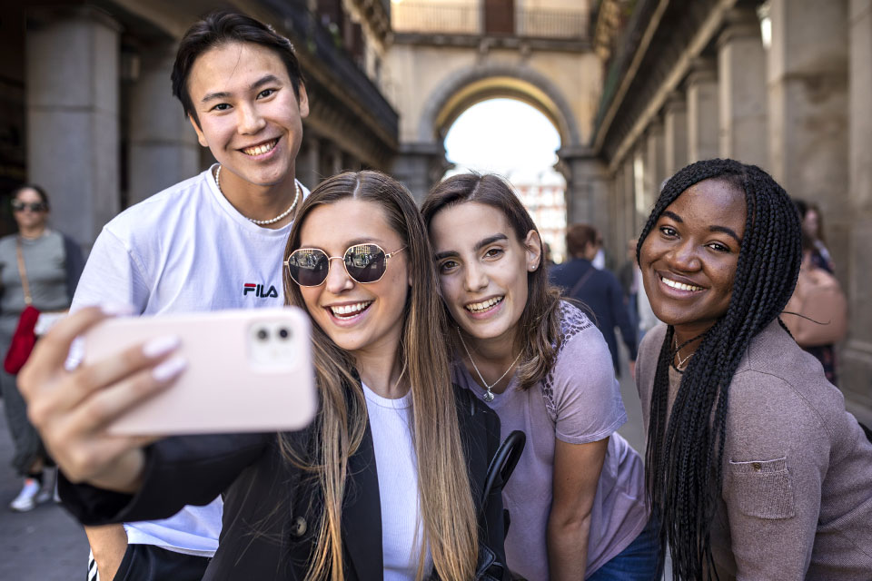 Four students smile while one holds a phone for a selfie in front of a stone structure in the city of Madrid.