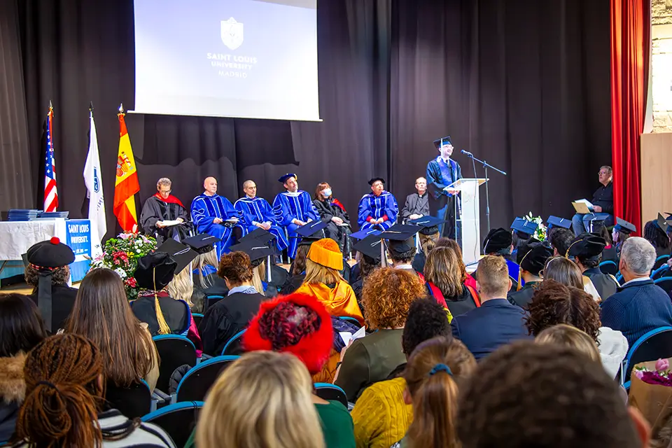 Audience members are shown from the back as a speaker in graduation regalia stands at a lectern. Additional people in graduation regalia sit behind the speaker on the podium.