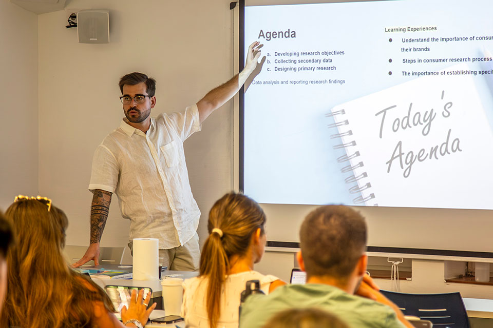 Miguel García faces a classroom of students while gesturing to a screen with a powerpoint slide that says Agenda.