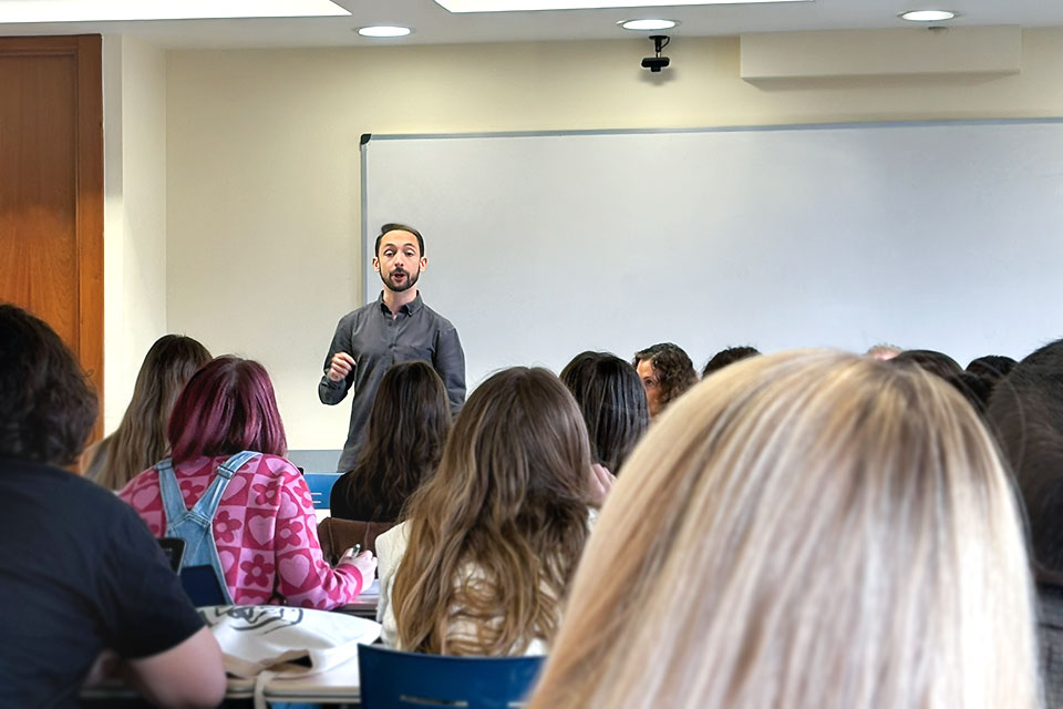 Students are seen from the back of their heads while a lecturer speaks at the front of a classroom.