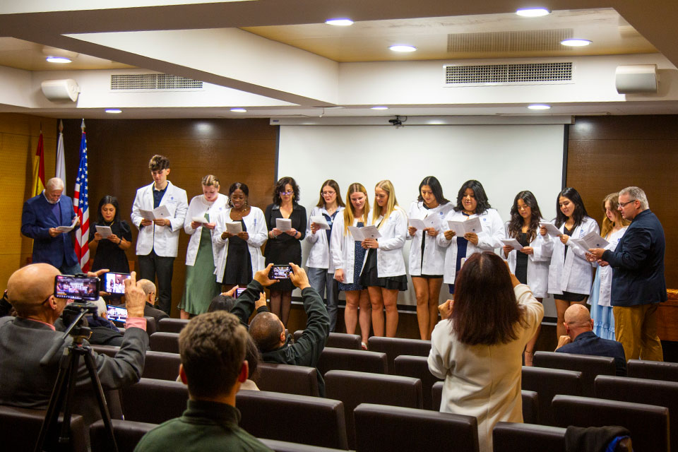 Students wearing white coats and faculty members read from pieces of paper at the front of an auditorium.