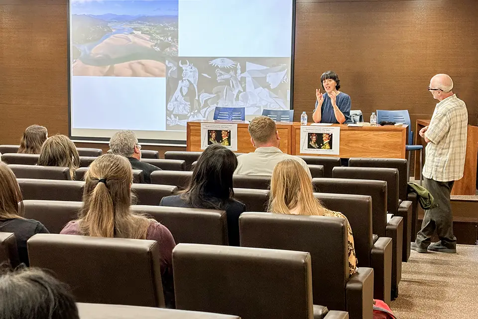Una autora sentada presenta su obra a una audiencia de estudiantes y profesores en un auditorio. / An author sitting down presents her work to an audience of students and professors in an auditorium.