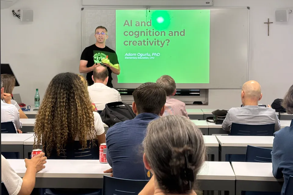 Man stands in front of people sitting at desks with a presentation projected onto a whiteboard behind him. The whiteboard reads AI cognition and creativity?