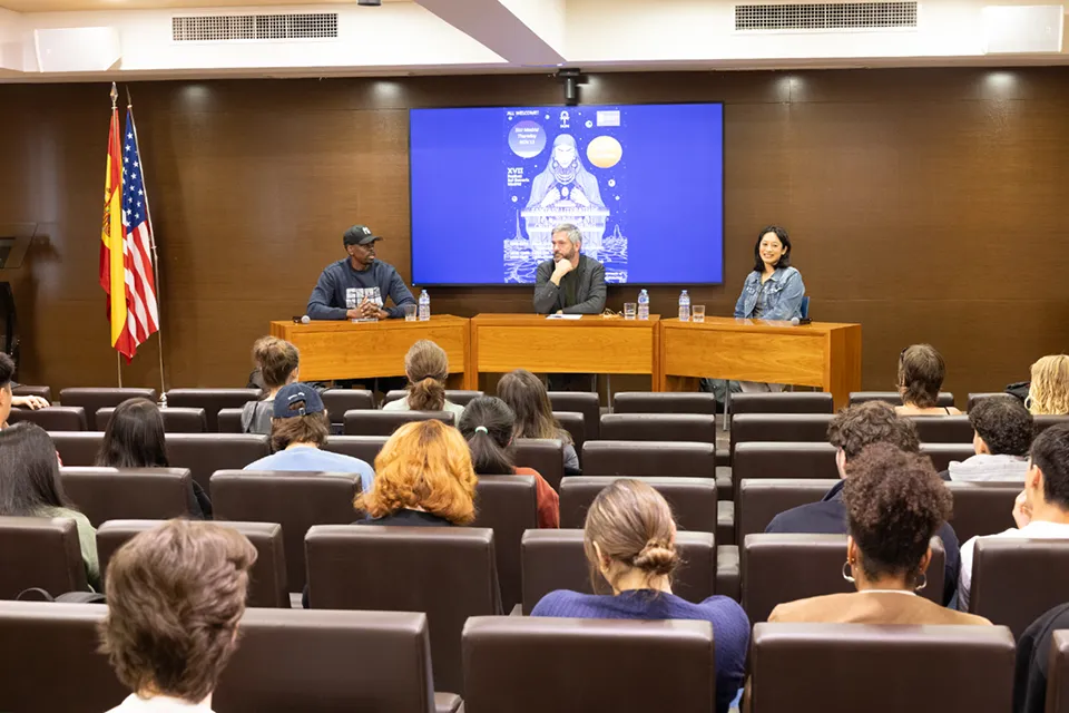 A panel of three speakers sits at a wooden table in front of an audience in a lecture hall. Behind them is a large screen displaying event artwork. The speaker on the left wears a cap and sweatshirt, the middle speaker rests his hand on his chin, and the speaker on the right smiles.