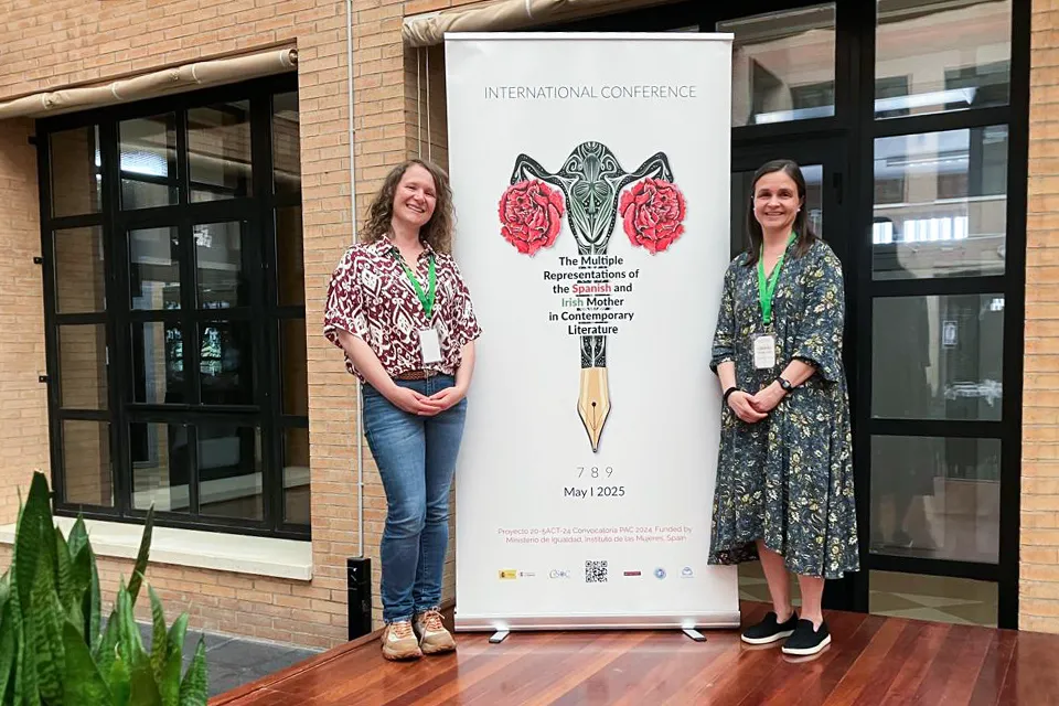 Two women wearing conference name tags stand on each side of a promotional poster for an international literature conference in the university patio. 