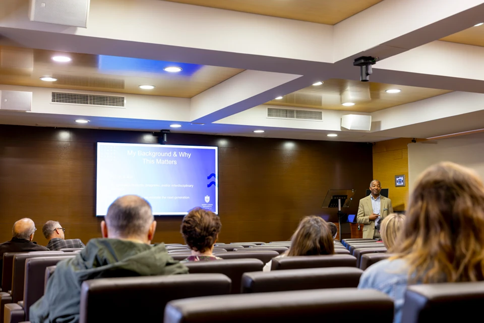 A man with a suit on looks at audience members who are sitting in a small auditorium with a screen that reads "My Background and Why This Matters."