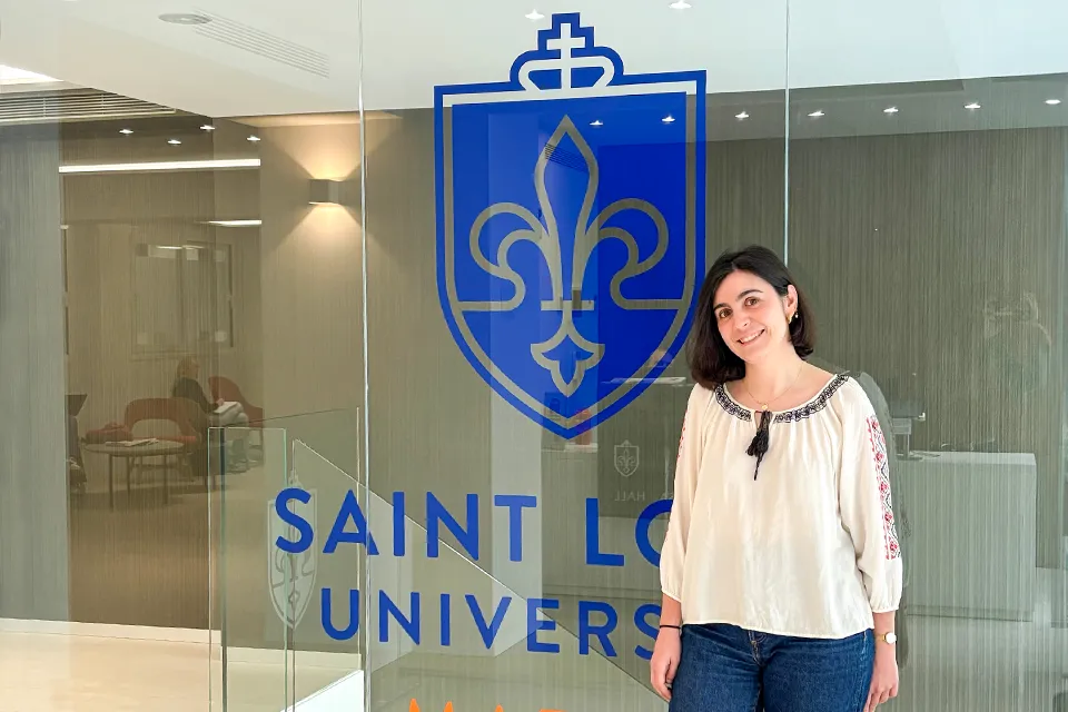 Woman looks at camera for picture with glass wall with university logo behind her.
