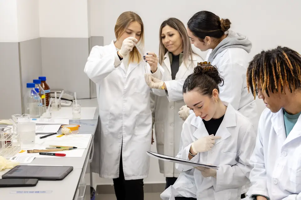 Chemistry class with professor Silvia Medina-Villar, Ph.D. Student holding specimen speaks with professor and fellow student while two other students look at notebook in a science lab.