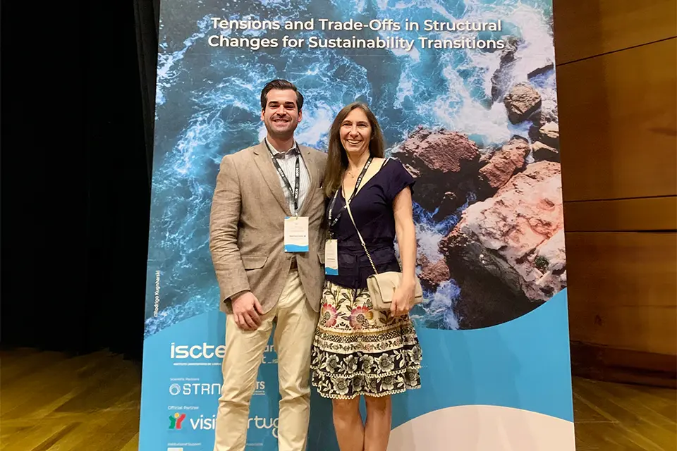 Two people stand and smile for picture with event poster in the background. 