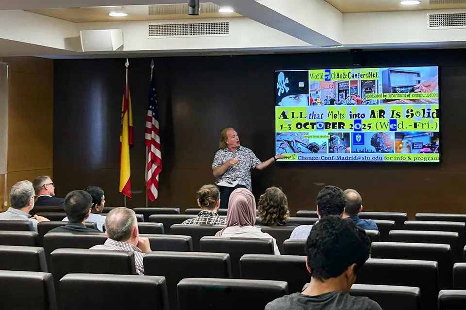 A speaker talks at the front of an auditorium in front of a video screen that reads "All that melts into air is solid"