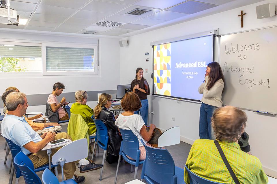 SLU-Madrid student volunteers Amelie and Gloria lead an advanced Community ESL class for Madrid residents. Two girls are standing next to the screen, teaching an English class, while the students listen to the lesson.