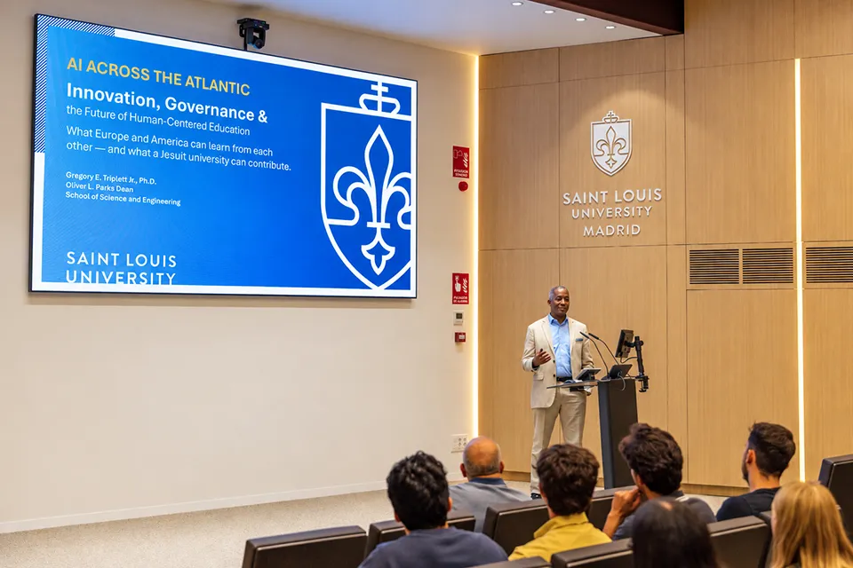 A man addresses the audience while a presentation titled “AI Across the Atlantic” is shown on a screen.