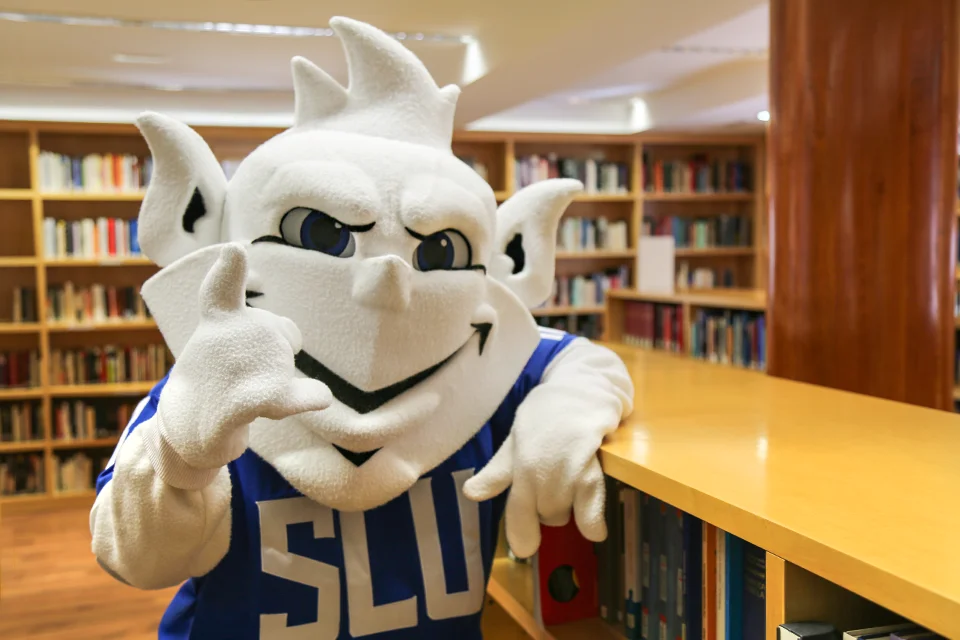 The Billiken visiting the SLU-Madrid Library in 2018. University mascot poses on library shelves with books in background.
