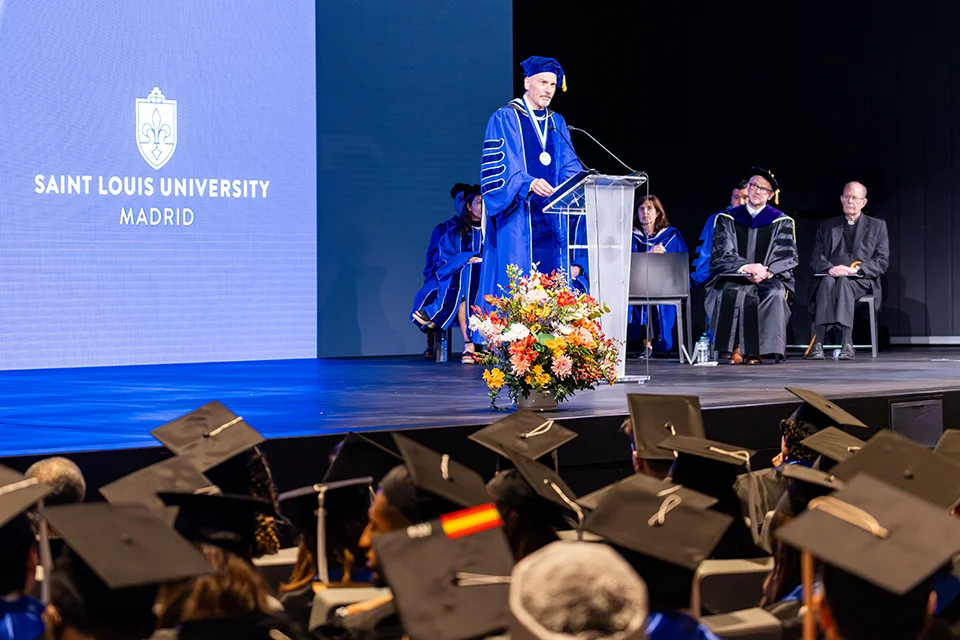 A man dressed in academic regalia speaks to the audience from a podium with stage party behind. In the background there is a screen where the University logo is projected. 
