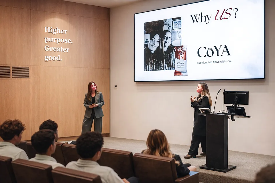 Two women in business attire speak to the audience with a presentation slide that shows the company logo. The wall of the auditorium has a sign that reads "Higher purpose. Greater good" in white.