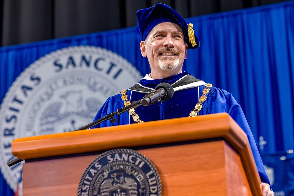 Man smiles looking to his left in academic regalia with a cap on that has a tassel. He is standing behind the podium that has the University seal on it. Behind him is a blue backdrop that also has the University seal on it.