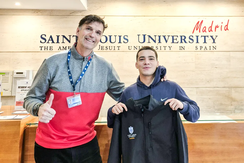 Man to the left gives a thumbs up and smiles at camera and student to his right looks on at camera while holding a sweater with the university logo. There is the university logo on the wall in the background behind a reception desk.